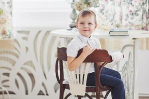 Small boy in blue trousers jeans is sitting on the wooden chair in the room photo