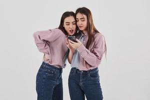 algún contenido interesante en sus teléfonos. compartiendo secretos. Dos hermanas gemelas de pie y posando en el estudio con fondo blanco.