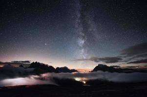 You can clear see the milky way. Something is shining in the middle of the forest with stars in the sky. The mountains are surrounded by dense fog photo