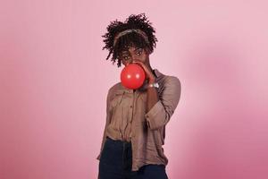 Portrait of young afro american girl blowing red balloon ar pink studio background photo