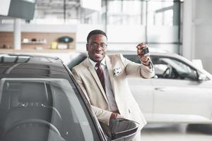 apuesto hombre negro en el concesionario está abrazando su nuevo coche y sonriendo foto