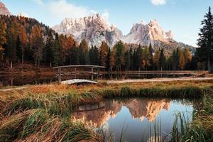 Sunlight on the top. Amazing view of majestic mountains with woods in front of them at autumn day. Puddle that goes from the lake with little bridge in the center photo