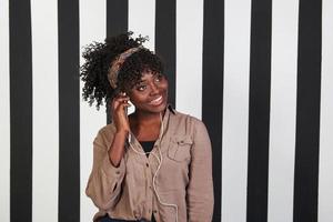 Putting headphone in ear and amazed my the music. Smiled afro american girl stands in the studio with vertical white and black lines at background photo