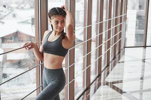 Touching the hair. Brunette girl in sportswear posing and laying on railings near window in the gym photo
