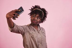 Yellow painted fingernails. Attractive afro american woman in casual clothes takes selfie at pink background in the studio photo