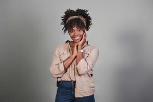 mujer positiva. Alegre niña afroamericana mantenga sus manos cerca de la cara mientras se encuentra en un fondo gris foto