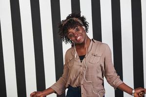 Woman listening music in headphones and dancing to it. Smiled afro american girl stands in the studio with vertical white and black lines at background photo