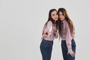 Looking straight into the camera and showing gestures. Two sisters twins standing and posing in the studio with white background photo