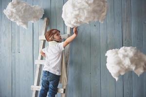 niño con sombrero de piloto de pie en la escalera en el estudio. llegar al cielo. tocar el concepto de nubes foto