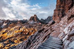 Sun lights going through the clouds. Extreme tourist path at the edge of the hills with wooden little bridge to the way to building standing at dolomites mountains photo