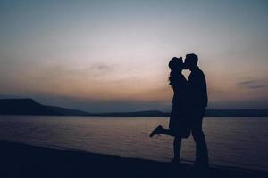 Siluetas de pareja besándose en el fondo del atardecer, el lago y las montañas