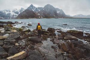 Traveler man with a yellow backpack wearing a red hat standing on the background of mountains and sea. Travel lifestyle concept. Shoot from the back photo