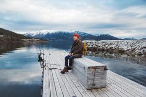 Traveler man with a yellow backpack wearing a red hat sitting on wooden pier on the background of mountain and lake. Space for your text message or promotional content