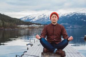 Traveler man in a meditative position sitting on a wooden pier on the background of a mountain and a lake