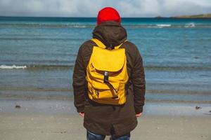 Traveler man with a yellow backpack wearing a red hat standing on a sandy beach on the background of the sea. Shoot from the back photo