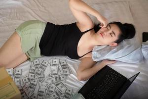 Portrait attractive young Asia woman sleeping on the bed in the bedroom with a laptop and stack 100 US dollars banknotes a lot of, works from own homes, Sell goods for profit concept photo