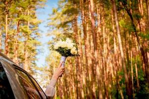 Bride hold white wedding bouquet out of cars window with scenic pine trees forest background . Wedding celebration outdoors in nature photo