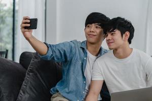 Romantic young gay couple funny selfie by cellphone at home. Asian lover male happy relax fun using technology mobile phone smiling take a photo together while lying sofa in living room concept.