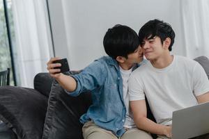Romantic young gay couple funny selfie by cellphone at home. Asian lover male happy relax fun using technology mobile phone smiling take a photo together while lying sofa in living room concept.