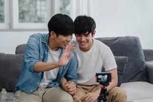 Young Asian gay couple influencer couple vlog at home. Teen korean LGBTQ men happy relax fun using camera record vlog video upload in social media while lying sofa in living room at house concept. photo