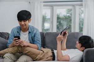 Young gay lgbtq couple using mobile phone and tablet at modern home. Asian lover male happy relax laugh and fun technology play games in internet together while lying sofa in living room concept. photo