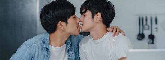 Young Gay couple kissing while using computer laptop at modern home. Asian LGBTQ men happy relax fun using technology play social media together while sitting table in kitchen at house concept. photo