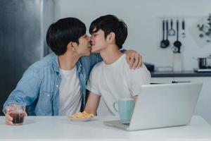 joven pareja gay besándose mientras usa computadora portátil en casa moderna. los hombres asiáticos lgbtq felices se relajan y se divierten usando la tecnología, juegan juntos en las redes sociales mientras se sientan a la mesa en la cocina en el concepto de casa. foto
