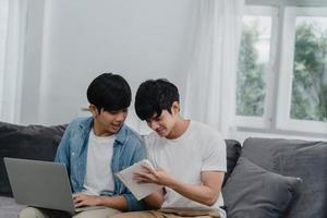 Young Asian Gay couple working laptop at modern home. Asia LGBTQ men happy relax fun using computer and analyzing their finances in internet together while lying sofa in living room at house concept.