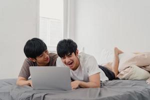 Asian Gay lgbtq men couple using computer laptop at modern home. Young Asia lover male happy relax rest together after wake up, watching movie lying on bed in bedroom at house in the morning concept. photo