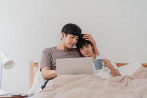 Asian Gay men couple using computer laptop and drinking coffee at modern home. Young Asia lover male happy relax rest together after wake up, watching movie lying on bed in bedroom at house in morning photo