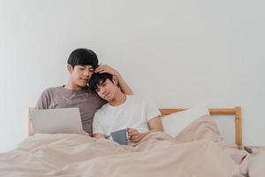 Asian Gay men couple using computer laptop and drinking coffee at modern home. Young Asia lover male happy relax rest together after wake up, watching movie lying on bed in bedroom at house in morning photo