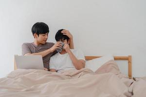 Asian Gay men couple using computer laptop and drinking coffee at modern home. Young Asia lover male happy relax rest together after wake up, watching movie lying on bed in bedroom at house in morning photo