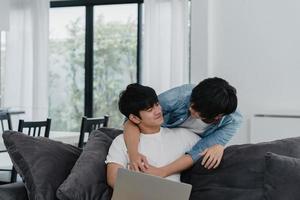Young Gay couple using computer laptop at modern home. Asian LGBTQ men happy relax fun using technology watching movie in internet together while lying sofa in living room at house concept. photo