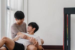 Asian Gay couple lying and hugging on the floor at home. Young Asian LGBTQ men kissing happy relax rest together spend romantic time in living room with rainbow flag at modern house in the morning. photo