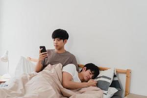 Asian Gay couple using mobile phone at home. Young Asia LGBTQ man happy relax rest after wake up, check social media while his boyfriend sleep lying on bed in bedroom at home in the morning concept. photo