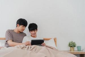 Asian Gay couple using tablet at home. Young Asian LGBTQ men happy relax rest together after wake up, check mail and social media lying on bed in bedroom at home in the morning concept. photo