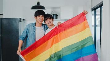 Retrato joven pareja gay asiática que se siente feliz mostrando la bandera del arco iris en casa. Los hombres lgbtq de Asia relajan una sonrisa con dientes mirando a la cámara mientras se abrazan en la cocina moderna de la casa en el concepto de la mañana. foto