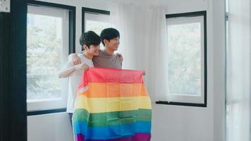 Retrato joven pareja gay asiática que se siente feliz mostrando la bandera del arco iris en casa. Los hombres lgbtq de Asia relajan una sonrisa con dientes mirando a la cámara mientras se abrazan en la moderna sala de estar de la casa en el concepto de la mañana. foto
