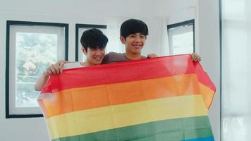 Retrato joven pareja gay asiática que se siente feliz mostrando la bandera del arco iris en casa. Los hombres lgbtq de Asia relajan una sonrisa con dientes mirando a la cámara mientras se abrazan en la moderna sala de estar de la casa en el concepto de la mañana. foto