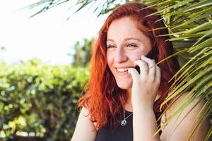 Close up of smiling attractive girl with redhead talking on mobile phone while standing outdoors photo