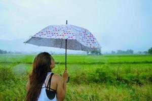 las mujeres asiáticas viajan relajarse en las vacaciones. la mujer de pie sostiene un paraguas bajo la lluvia feliz y disfrutando de la lluvia que cae. viajando en campiña, campos de arroz verde, viaje a tailandia. foto