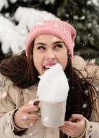 woman in warm winter clothes standing by the big christmas tree outdoors and licking snow from the cup photo