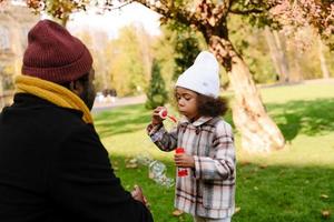 Black girl blowing soap bubbles while walking with grandfather in an autumn park