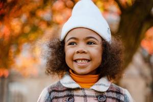 Chica rizada negra con sombrero blanco sonriendo mientras camina en el parque de otoño