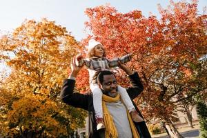 Chica negra divirtiéndose y sentada en el cuello de su abuelo en el parque de otoño