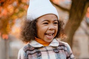 Chica rizada negra con sombrero blanco sonriendo mientras camina en el parque de otoño