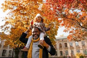 Chica negra divirtiéndose y sentada en el cuello de su abuelo en el parque de otoño foto