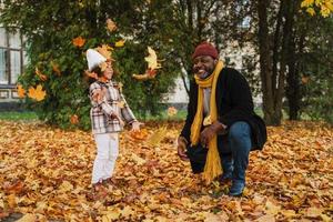 Abuelo negro y nieta burlándose con hojas caídas en el parque de otoño