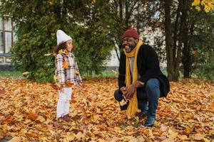 Abuelo negro y nieta burlándose con hojas caídas en el parque de otoño