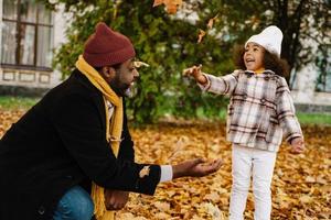 Black grandfather and granddaughter having fun with fallen leaves in autumn park photo
